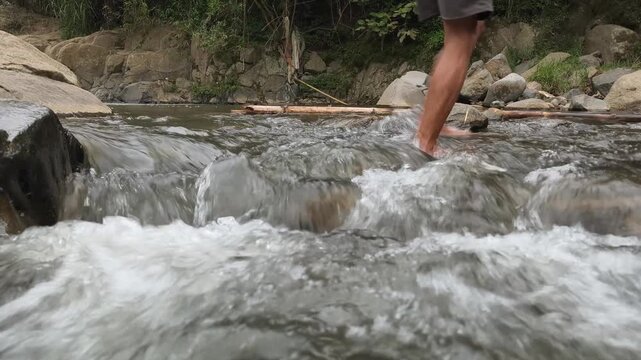 A man walks across a river or wild stream barefoot. Steady shot of a clear, fast-flowing river with bare feet crossing.