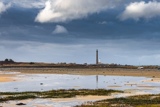 View of Ile Vierge lighthouse standing tall on a rocky island across a sandy beach at low tide under a dramatic cloudy sky Plouguerneau, Bretagne, France.