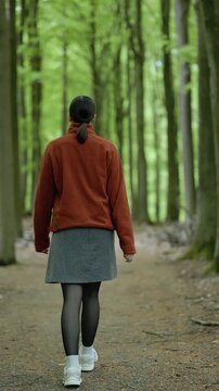 Back view of a young woman in a bright orange sweater and skirt walking along a forest path and stopping to breathe fresh air in a peaceful natural environment.