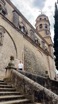 A woman strolls along ancient stone steps beside a grand Renaissance cathedral with an ornate bell tower. Dramatic low angle view emphasizes the imposing sandstone architecture against a cloudy sky. 