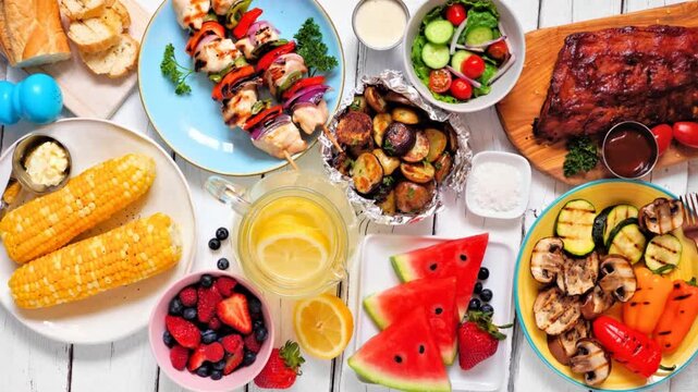 Summer BBQ or picnic food table scene. Slow rotating zoom motion. Assortment of grilled meats, vegetables, fruits, salad and potatoes. Top down view on a white wood background.