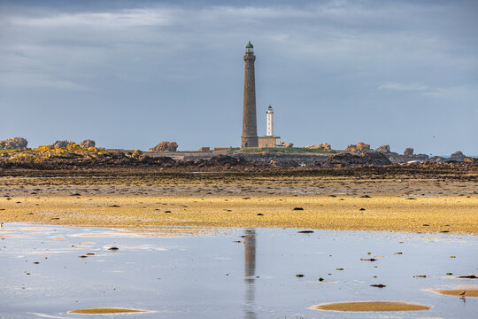 View of the Ile Vierge lighthouse standing tall on a rocky islet with its reflection in the shallow tide pools of a sandy beach under a dramatic cloudy sky Plouguerneau, Bretagne, France.