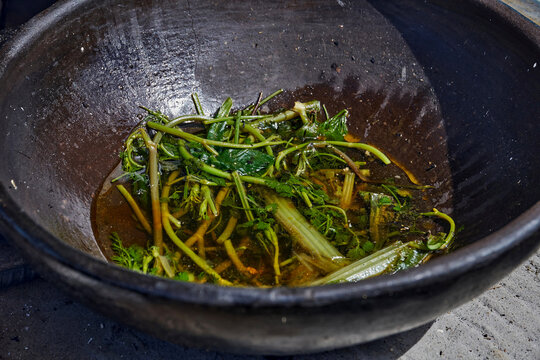 Herbal mixture of aromatic plants soaking in oil inside a large black bowl on textured surface, process of preparing an aromatic herbal base for traditional Ecuadorian dish in clay pot (olla de barro)