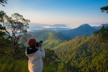 A rear-view shot of a female photographer wearing a white fleece jacket and an orange beanie, capturing a stunning mountain landscape with a professional camera. The scene overlooks lush green hills  © Hathaichanok