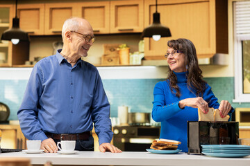 Fototapeta premium Senior couple preparing breakfast together in a modern kitchen, cozy morning routine. Husband and wife sharing a candid moment at kitchen counter, serving healthy food and warm coffee.