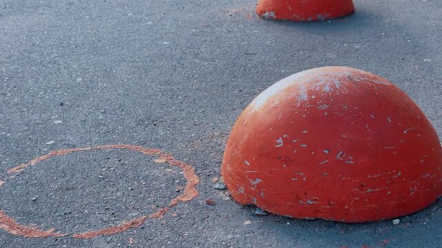 Red hemispheres on asphalt pedestrian path, row of weathered orange bollards creating rhythmic barrier in empty urban pedestrian