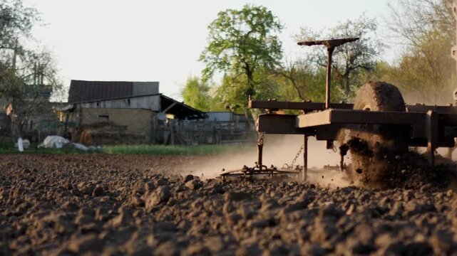Close-up cinematic shot of a blue tractor driving through a field during spring cultivation. The wheels and harrow move the soil, creating dust in the bright sunlight. Traditional agricultural machine