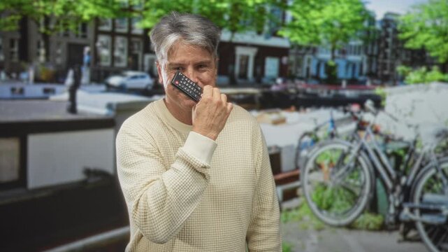 Man holding tv remote to cheek on a street canal with parked bicycles and buildings; contentment leisure.