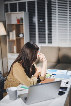 Asian businesswoman having a stressful phone call, holding a pen, and looking troubled while working on her laptop with financial documents and a calculator on her office desk