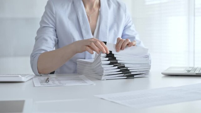 Businesswoman's hands organizing a towering stack of bound reports and financial documents on a white desk, navigating paperwork overload in a busy corporate office environment. Audit and taxes