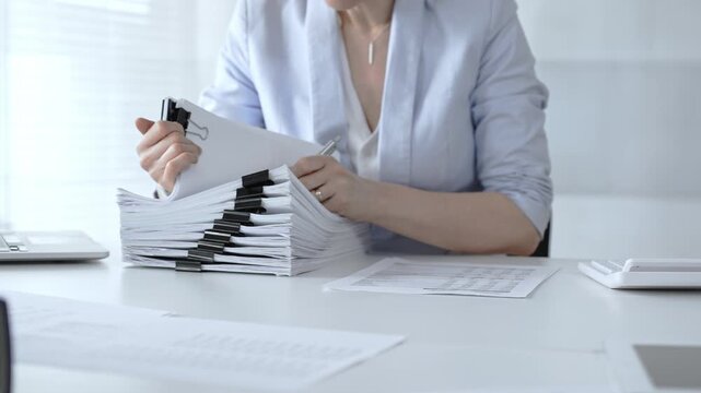 Businesswoman's hands organizing a towering stack of bound reports and financial documents on a white desk, navigating paperwork overload in a busy corporate office environment. Audit and taxes
