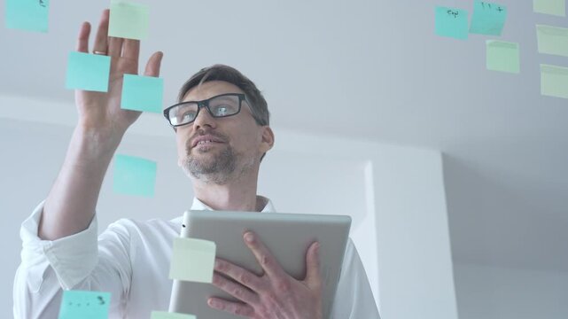 Adult businessman in glasses using a tablet computer while brainstorming and organizing work on a transparent glass board with colorful sticky notes, focusing on strategy and planning