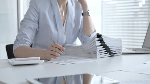 Businesswoman's hands organizing a towering stack of bound reports and financial documents on a white desk, navigating paperwork overload in a busy corporate office environment. Audit and taxes