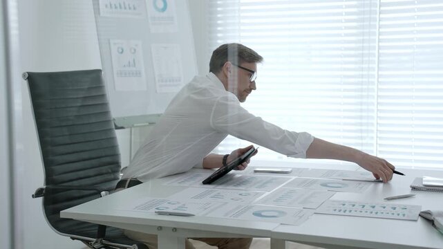 Businessman in modern office studying charts and reports at desk, concentrating on market data and strategic planning. Business concept