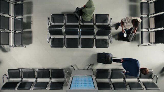 Top down view of crowd walking in busy airport next to FIDS electronic display. Passengers navigating transit hub during long flight layover, passing by departure board display, high angle shot