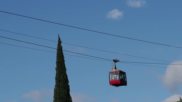 red funicular car leading to the top of the old mountain. The tourist car rides above rocky mountains dotted with cypress trees. Cable car cabins amazing sky and clouds. Cableway, green transportation