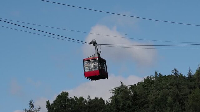 red funicular car leading to the top of the old mountain. The tourist car rides above rocky mountains dotted with cypress trees. Cable car cabins amazing sky and clouds. Cableway, green transportation