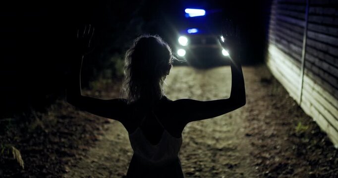 Girl stands with hands raised in front of police silhouette at night on a deserted road with a flashing light