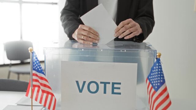 Woman placing ballot into clear ballot box showing hands and ballot at a voting building; civic participation determination.