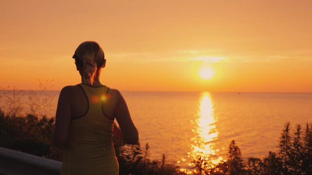 Silhouette of woman jogging along coastal road at sunset with golden light reflecting on water surface during evening cardio workout in slow motion representing healthy lifestyle and fitness goals.