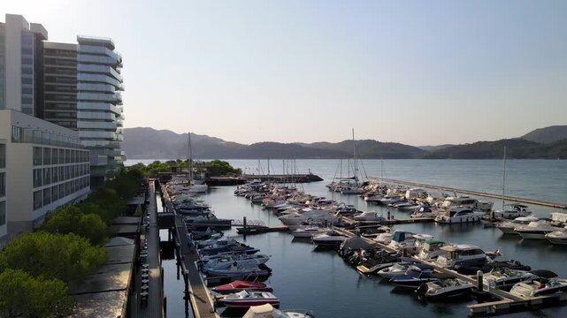 Cinematic drone shot flying at low altitude over parked sailboats and motorboats in Troia Marina. The footage captures the golden hour light over the harbor, modern waterfront architecture, and the tr