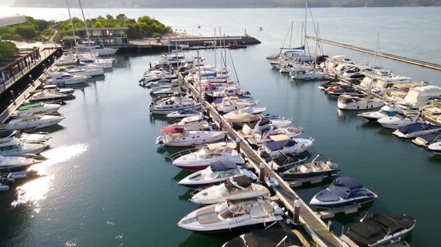 Cinematic drone shot flying at low altitude over parked sailboats and motorboats in Troia Marina. The footage captures the golden hour light over the harbor, modern waterfront architecture, and the tr