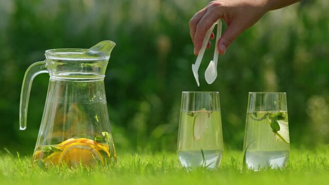 Fresh lemonade preparation on green grass with glass pitcher and two tumblers filled with citrus mint water while female hand adds lime slice using ice tongs during sunny summer garden party event.
