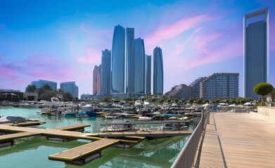 Naklejka premium Iconic Abu Dhabi waterfront skyscrapers tower above a bustling marina filled with luxury yachts, viewed from a wooden boardwalk promenade at a colorful sunset, UAE