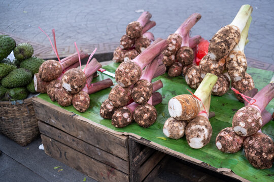 Fresh talas (taro roots) with purple stalks arranged on a wooden market table in Bogor, beside a basket of soursop fruits, showing freshly harvested local produce for daily trade.