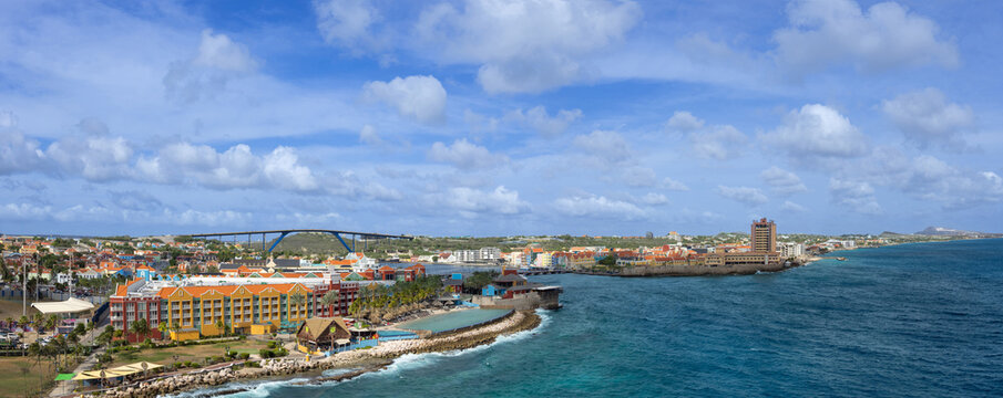 A wide panoramic view of the Willemstad waterfront in Curacao featuring colorful colonial buildings along the coast a luxury resort Queen Juliana Bridge and the blue Caribbean sea