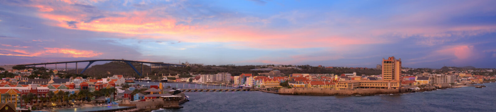 A wide sunset panoramic view of the Willemstad waterfront in Curacao featuring colorful colonial buildings along the coast a luxury resort Queen Juliana Bridge and the blue Caribbean sea