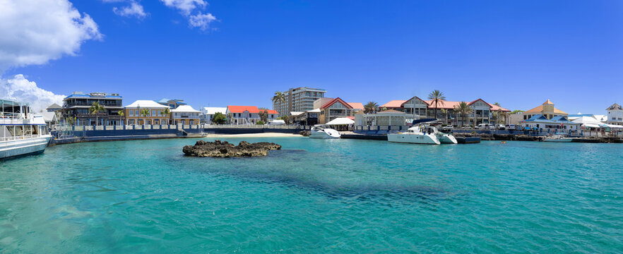 Panoramic view of the George Town waterfront in Grand Cayman featuring colorful commercial buildings and white boats on crystal clear turquoise Caribbean water under a bright blue cloudy sky