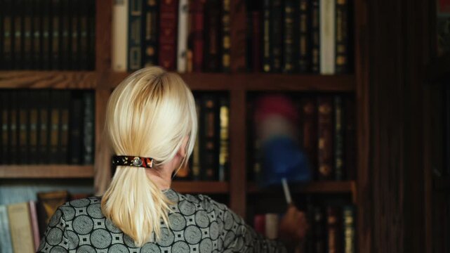 Blonde woman with a ponytail cleaning dust from books on a wooden bookshelf in a library using a colorful duster, showcasing diligent motion and domestic care in her routine duties.