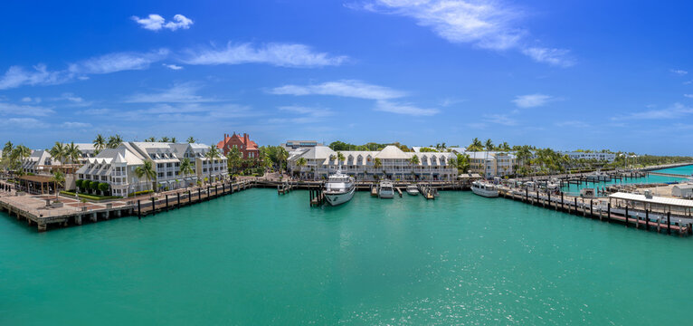 Panoramic view of Key West harbor, Florida, featuring a marina with docked boats, white waterfront buildings, palm trees and vivid turquoise water under a bright blue sky