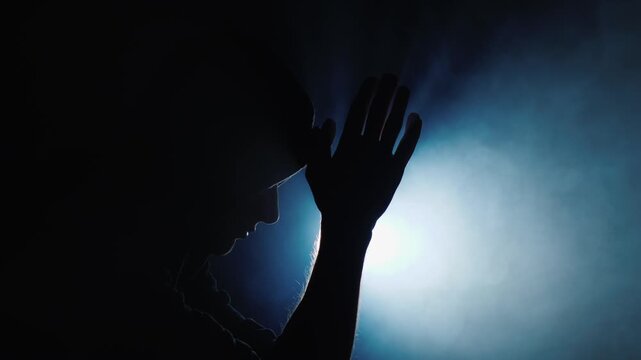 Silhouette of man in profile with hands together in prayer position on dark blue background with soft dramatic light and smoke effects cinematic motion footage highlighting spiritual contemplation.