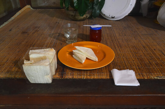 A top-down perspective showing slices of white bread on a plate, accompanied by a jar of red strawberry jam, a glass of water, and bread packaging on a kitchen table.