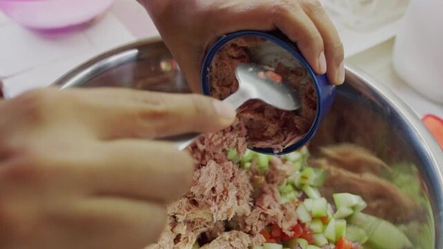 Close up of hands using a spoon to empty a can of tuna into a bowl of chopped vegetables. Preparing a healthy tuna ceviche or fish salad for a meal.