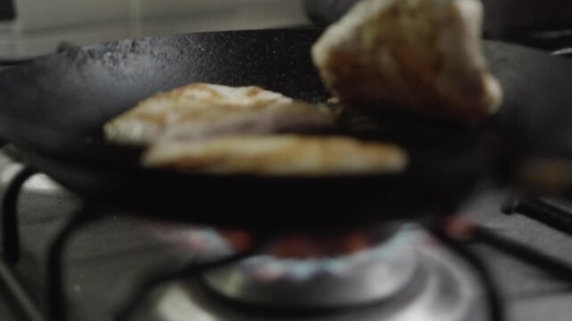 Chicken fillets cooking in black pan over visible gas flame, close up kitchen shot