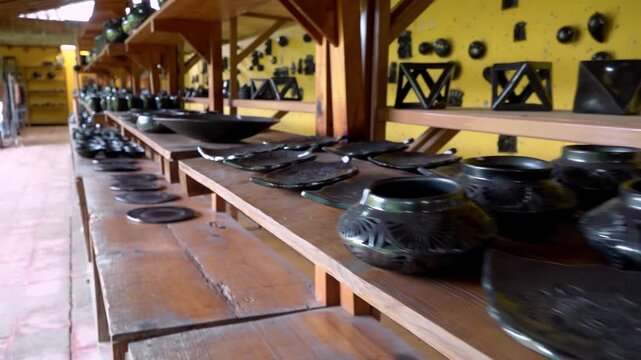 Row of handmade pottery arranged on wooden shelves inside artisan workshop, dark clay bowls and decorative items showing craft tradition, culture and rustic interior setup