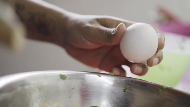 Closeup view of hand breaking egg above metal bowl with ingredients, step of adding egg to chicken meatball mixture, simple home cooking preparation in kitchen setting