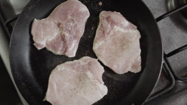 Birds eye view of raw pork loin fillets searing in a black frying pan on a kitchen stove. Topdown shot of meat preparation and home cooking.