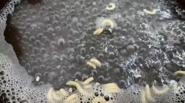 Close view of boiling liquid with floating noodle pieces, dense bubbles, foam clusters, and reflective ripples forming textured patterns across the surface in close detail clearly.