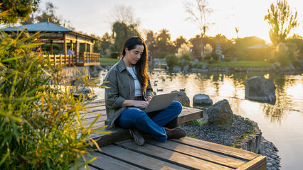 Woman typing on a laptop, enjoying outdoor office work by a tranquil lake at sunset © freechoice