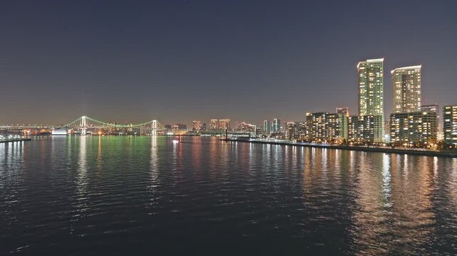 Wide static time-lapse of Tokyo Bay at night, Rainbow Bridge with green cable lights left of frame, tall glittering residential towers right, multicolored reflections shimmering on calm dark water.
