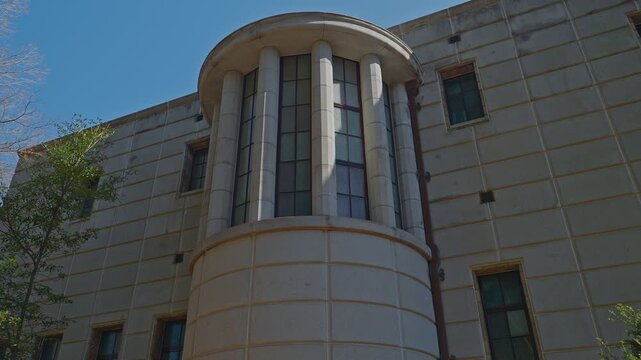 Low-angle static shot of a historic stone building's cylindrical corner tower with tall classical columns, multi-pane grid windows, strong sunlight casting sharp shadows against vivid blue sky.