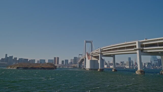 Rainbow Bridge's white suspension tower and curved ramp span a windy Tokyo Bay, with a wooded island and distant city skyline, captured as a static wide shot in bright daylight.