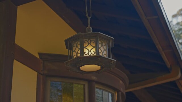 Close-up static shot of an antique wrought-iron hanging lantern glowing warmly beneath dark wooden beam ceiling, curved bay window and warm ochre wall visible in soft background.