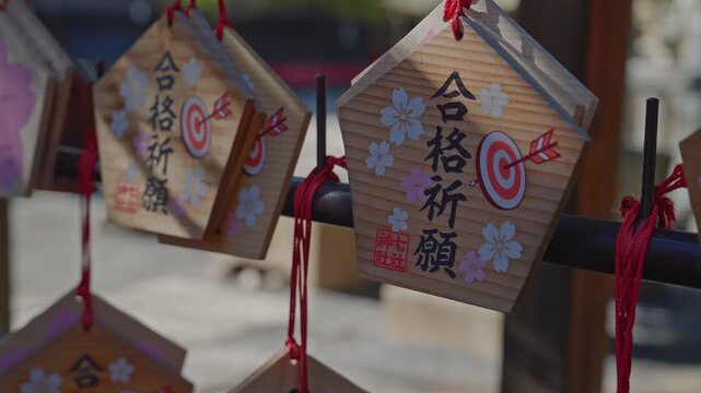Close-up static shot of hanging wooden ema prayer plaques at a Japanese shrine, painted with kanji wishing for exam success, archery target and arrow motifs, cherry blossom flowers, tied with red stri