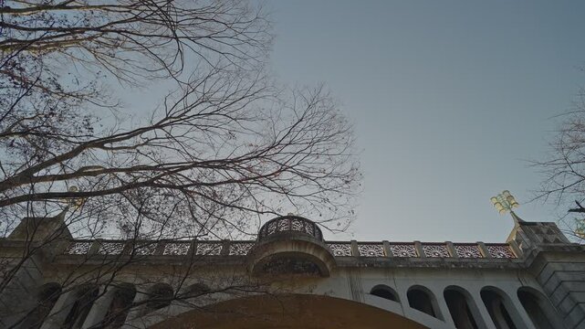 Ornate lattice parapet and curved balcony stretching across mid-frame, golden lamp post upper right, bare winter branches spreading against pale blue-grey dusk sky.