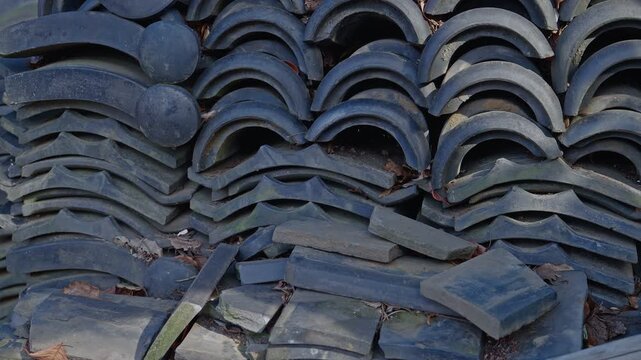 Close-up static shot of weathered dark grey traditional Japanese ceramic roof tiles stacked in irregular piles outdoors, curved and flat tile forms layered with dry leaves caught between, soft natural